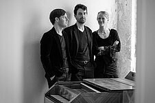 Schwarzweissfoto mit drei Personen, die hinter einem Cembalo stehen. Rechts der schmale Spalt eines Fensters..