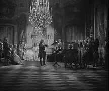 Schwarzweißfoto mit Konzertszene. Fußboden mit Rautenmuster, in der Mitte ein dunkel gekleideter Flötist, rechts davon ein Cembalist. Im Hintergrund weibliches und Männliches Publikum in barocke Kostümen