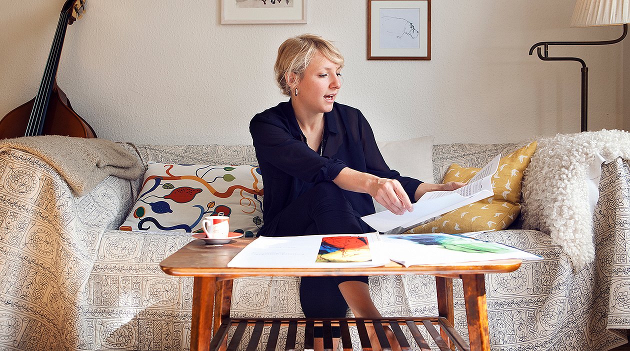 Eine blonde Frau sitzt auf einem blumigen Sofa, vor sich einen Couchtisch aus hellem Holz mit Notenblättern