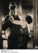 Viragiertes Foto mit einem Mann in Polizeiuniform und einer Frau mit Bubikopf in schulterfreiem Kleid, die sich an seinen Hals hängt.