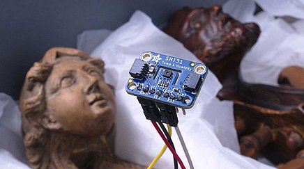 Elektronisches Messgerät im Vordergrund. Im Hintergrund liegen zwei hölzernen Figuren