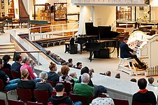 Mann sitzt an der Orgel und spielt. Publikum hört zu.