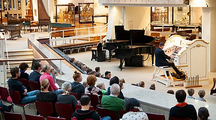 Mann sitzt an der Orgel und spielt. Publikum hört zu.