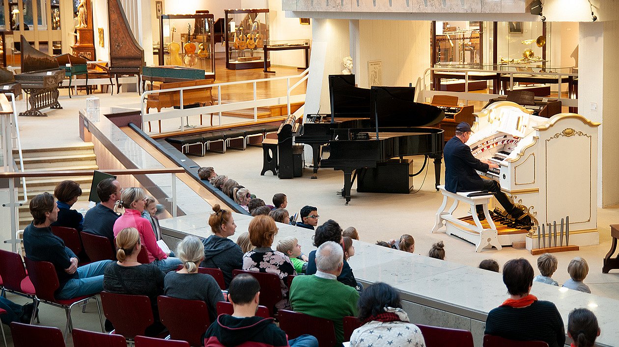Mann sitzt an der Orgel und spielt. Publikum hört zu.