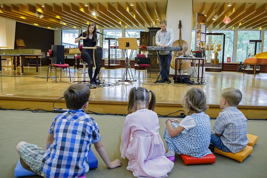 Vier Kinder sitzen vor einer Bühne, auf der eine Frau Theremin spielt und ein Mann Keyboard.