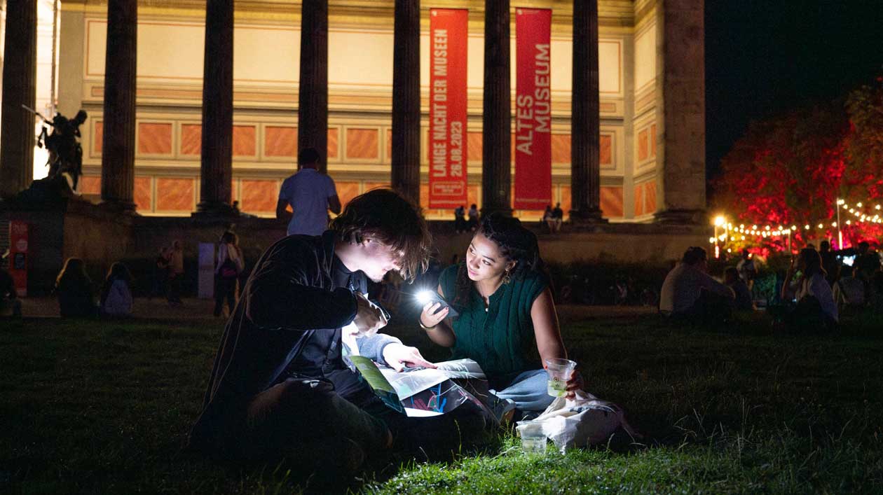 Zwei junge Personen sitzen an Abend vor einem prachtvollen Museum und beleuchten mit Taschenlampen ein Programmheft