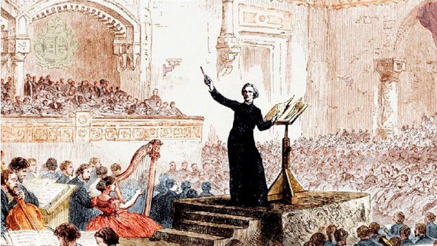 Ein Mann im schwarzen Gewand dirigiert ein Orchester. Sitzendes Publikum ist im Hintergrund zu sehen. 