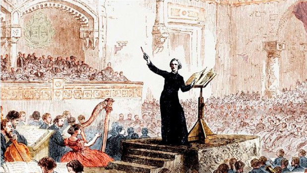 A man in a black robe conducts an orchestra. A seated audience can be seen in the background.