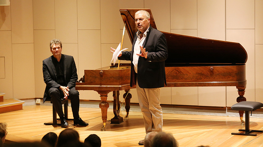 On a wooden stage: man at a grand piano made of shiny brown wood and man standing next to it talking.