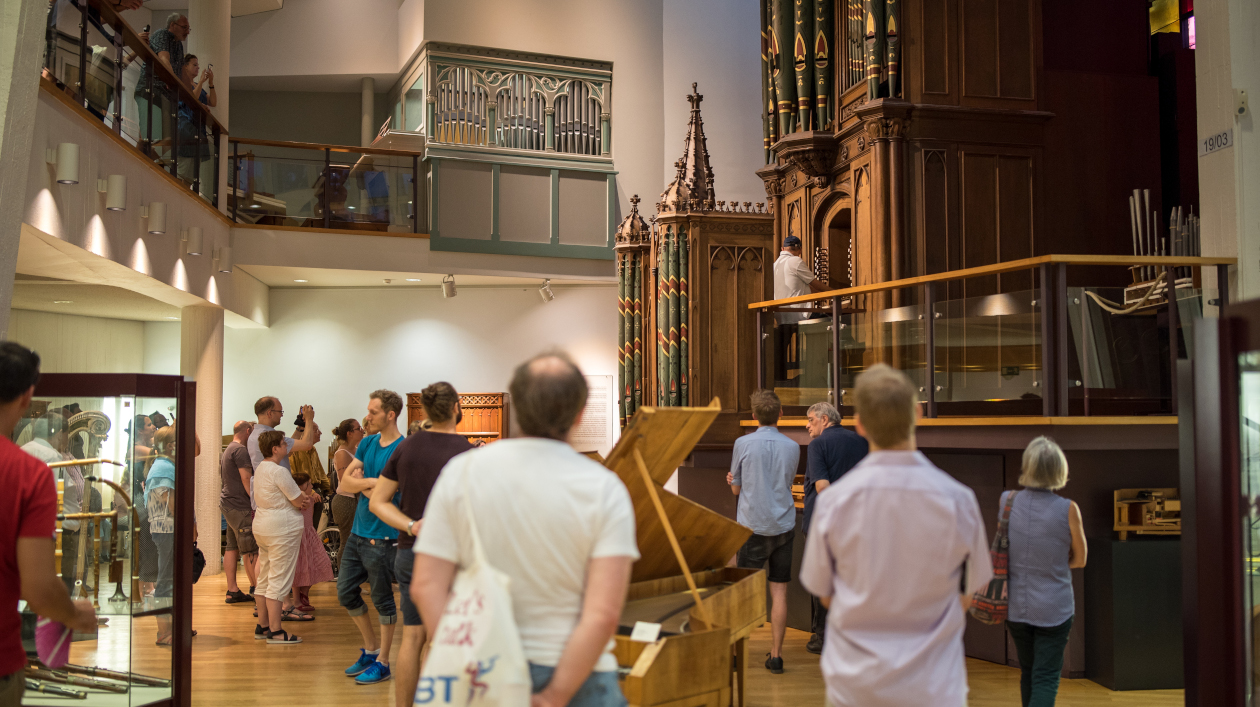 Besucher im Museum, die auf hellem Holzparkett stehend bei einer Präsentation der Gray-Orgel zuhören. Die Orgel im neugotischen Stil sieht man im Hintergrund. Im Vordergund stehen Vitrinen mit Instrumenten.