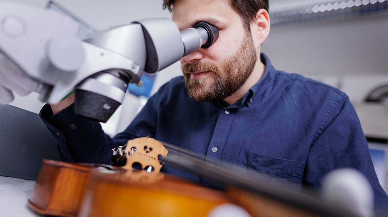 Man with beard in blue shirt looks into a microscope. In front of him is a violin.