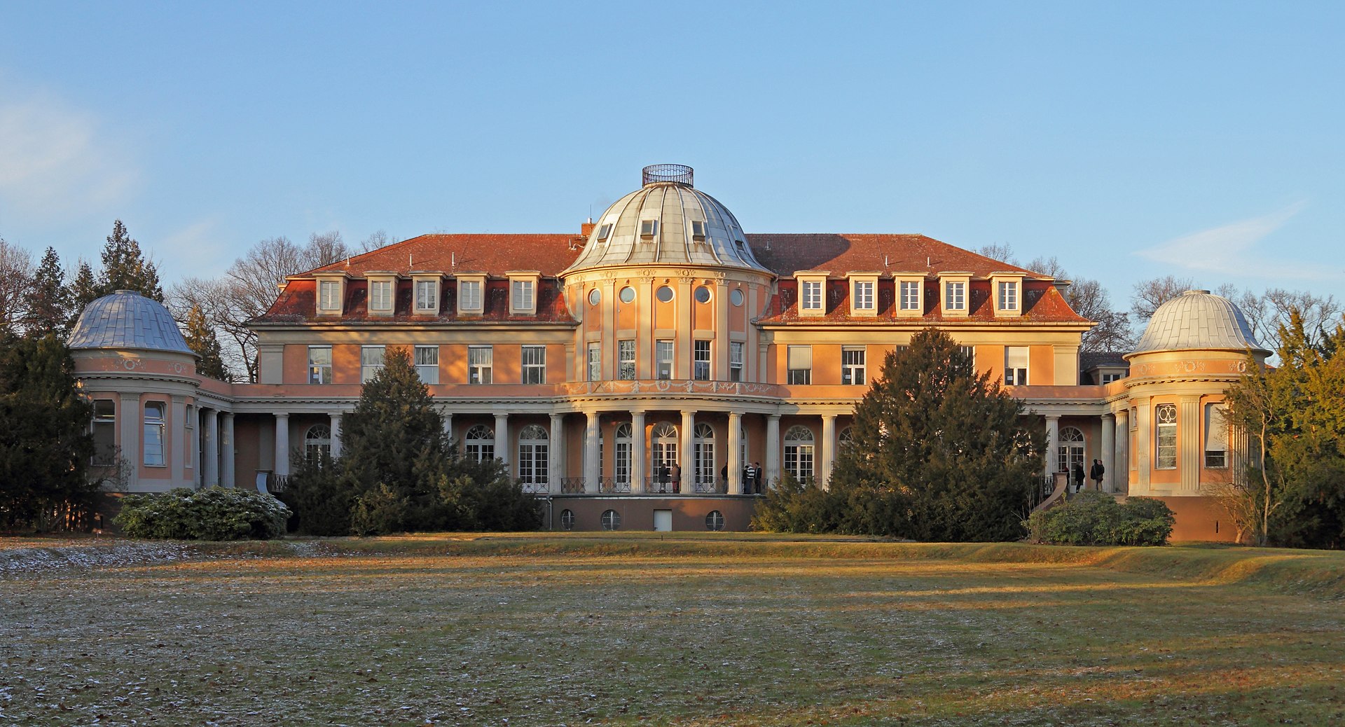 Siemensvilla in Berlin Lankwitz Blick aus dem Garten auf die Rückseite der lachsfarbenden Siemensvilla in Berlin Lankwitz. Heute wird das Gebäude Herrenhaus Correns genannt. In der Mitte und an den Seiten der Villa mit rotem Dach befinden sich Runderker, welche mit einem Säulengang verbunden sind.