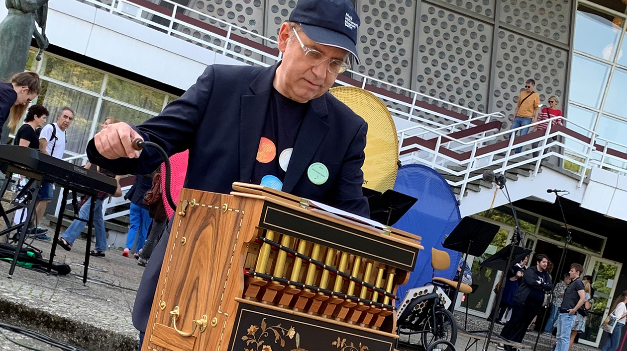 A man in a colourful shirt with a baseball cap plays a barrel organ. The Berlin Philharmonic Hall can be seen in the background.